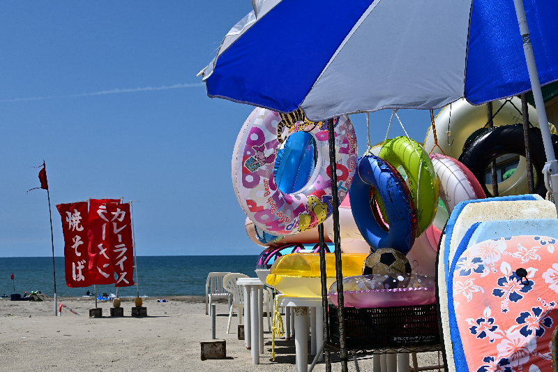 海をバックに海の家で浮き輪やサーフボードが積まれている画像